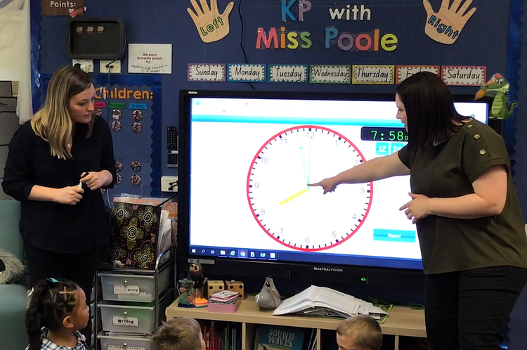 2 teachers standing at an interactive whiteboard and teaching a class about time.